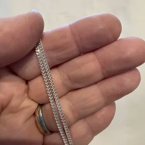 En hand håller i Silverkedja pansar 2.1mm halskedja (50-60cm) och visar dess geometriska, sammanlänkade silverkedjedesign. Personen bär också en guld- och silverring på sitt ringfinger, med en vanlig suddig bakgrund.