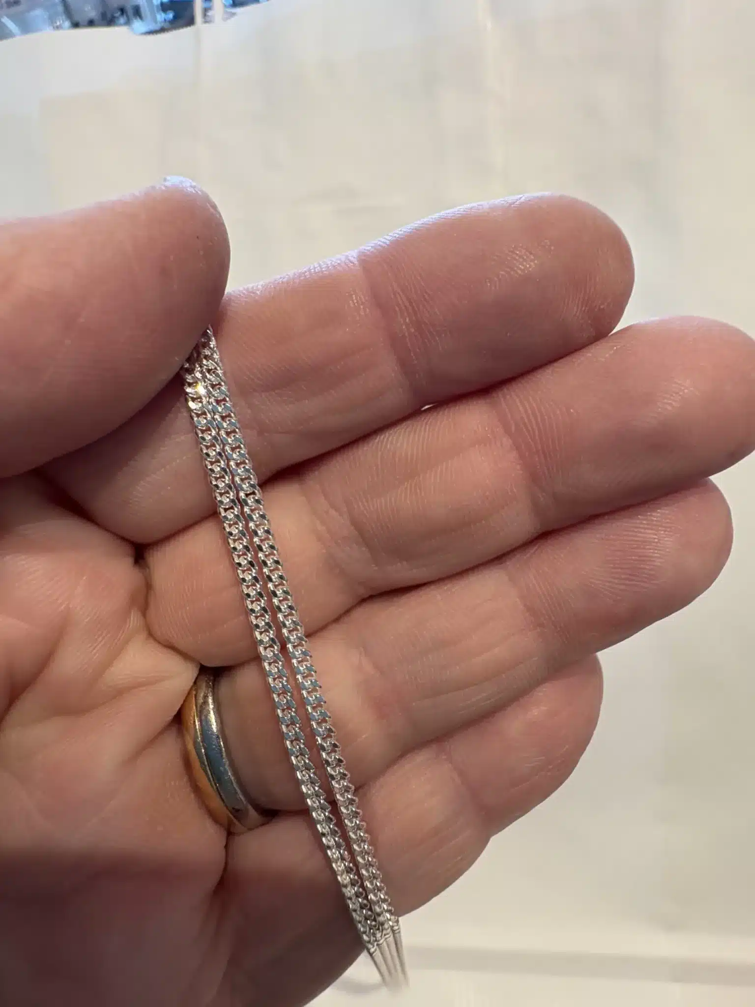 En hand håller i Silverkedja pansar 2.1mm halskedja (50-60cm) och visar dess geometriska, sammanlänkade silverkedjedesign. Personen bär också en guld- och silverring på sitt ringfinger, med en vanlig suddig bakgrund.