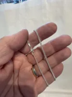 En hand håller i Silverkedja pansar 2.1mm halskedja (50-60cm), ett tunt silverhalsband med karbinlås; personen bär en vigselring i guld mot en suddig ljus bakgrund.