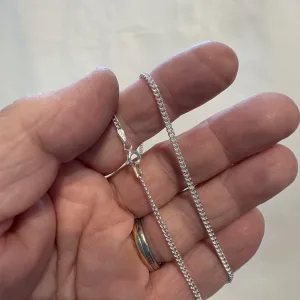 En hand håller i Silverkedja pansar 2.1mm halskedja (50-60cm), ett tunt silverhalsband med karbinlås; personen bär en vigselring i guld mot en suddig ljus bakgrund.