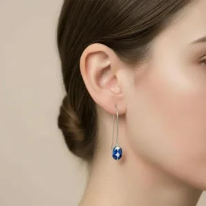 En närbild av en kvinnas öra visar Örhänge Oval - Adam: ett silverörhänge med en blå och vit mönstrad pärla, parat med hennes bruna hår i en låg knut.