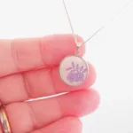 En hand håller i Porslinssmycke - Lila Fasan, ett silverhalsband med ett runt hänge som visar en enkel lila blomsterdesign. Bakgrunden är ljus och mjukt suddig.