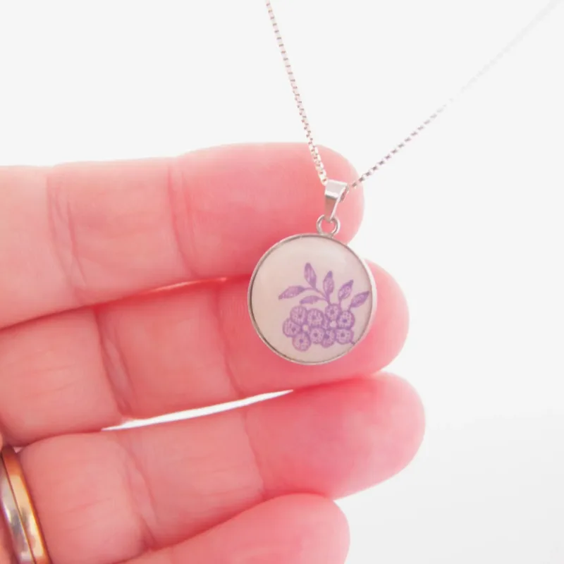 En hand håller i Porslinssmycke - Lila Fasan, ett silverhalsband med ett runt hänge som visar en enkel lila blomsterdesign. Bakgrunden är ljus och mjukt suddig.