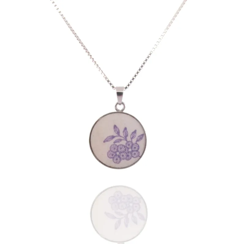 Porslinssmycke - Lila Fasan är ett silverhalsband med ett runt hänge prytt med ett fint lila blomster- och bladmotiv, mot en vit bakgrund med hängets reflektion synlig nedanför.