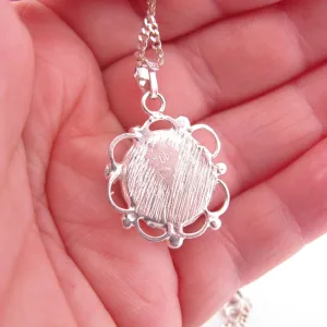 Närbild av en hand som håller Halsband med handslipad Hoburgsmarmor 32x23mm, med ett runt, texturerat marmorhänge med en utsmyckad skålad kant på en delikat silverkedja.