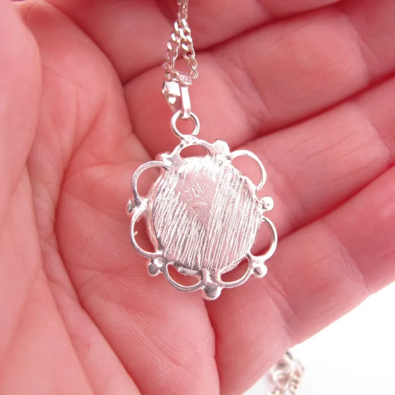 Närbild av en hand som håller Halsband med handslipad Hoburgsmarmor 32x23mm, med ett runt, texturerat marmorhänge med en utsmyckad skålad kant på en delikat silverkedja.