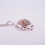 Halsband med handslipad Hoburgsmarmor 32x23mm" är ett silverhalsband med en rund, marmorerad Hoburgsmarmorsten i vitt, orange och grått, infattad i en utsmyckad infattning med dekorativa öglor och silverpärlor.
