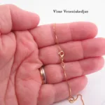 En hand med guldring håller i Hängsmycke i 14K Guld Gotland, ett delikat guldhänge med ett fint spänne. Bakgrunden är ljus och den svenska texten "Visar Veneziakedjan" syns i det övre högra hörnet.