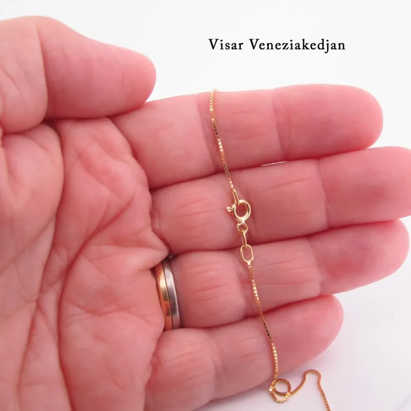 En hand med guldring håller i Hängsmycke i 14K Guld Gotland, ett delikat guldhänge med ett fint spänne. Bakgrunden är ljus och den svenska texten "Visar Veneziakedjan" syns i det övre högra hörnet.