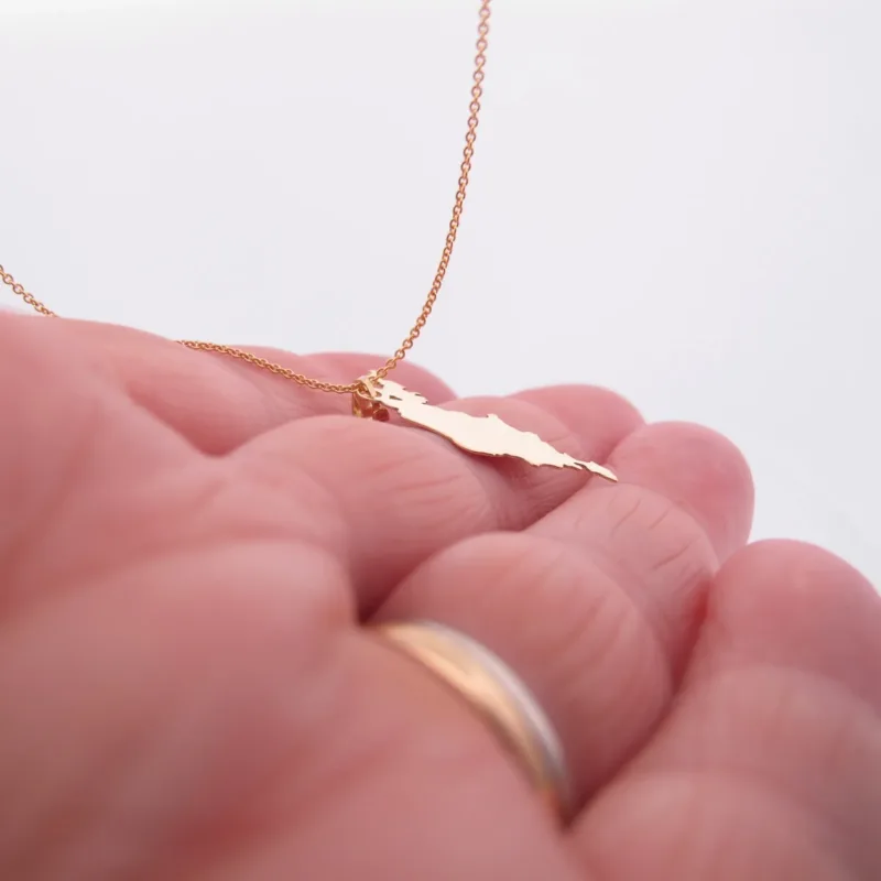 En hand håller i Hängsmycke i 14K Guld Gotland - ett guldsmycke format som Gotland - med en vigselring på ett finger. Den mjuka, suddiga bakgrunden framhäver den eleganta designen.