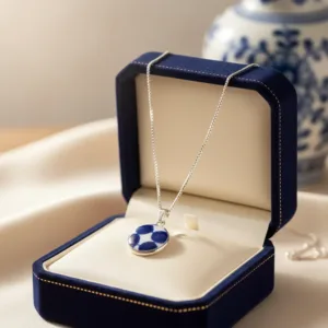 Porslinssmycke Adam 22x15mm silverhalsband med ett blått och vitt porslinshänge i en öppen smyckeskrin i marinblå sammet, med en suddig keramikvas i bakgrunden.