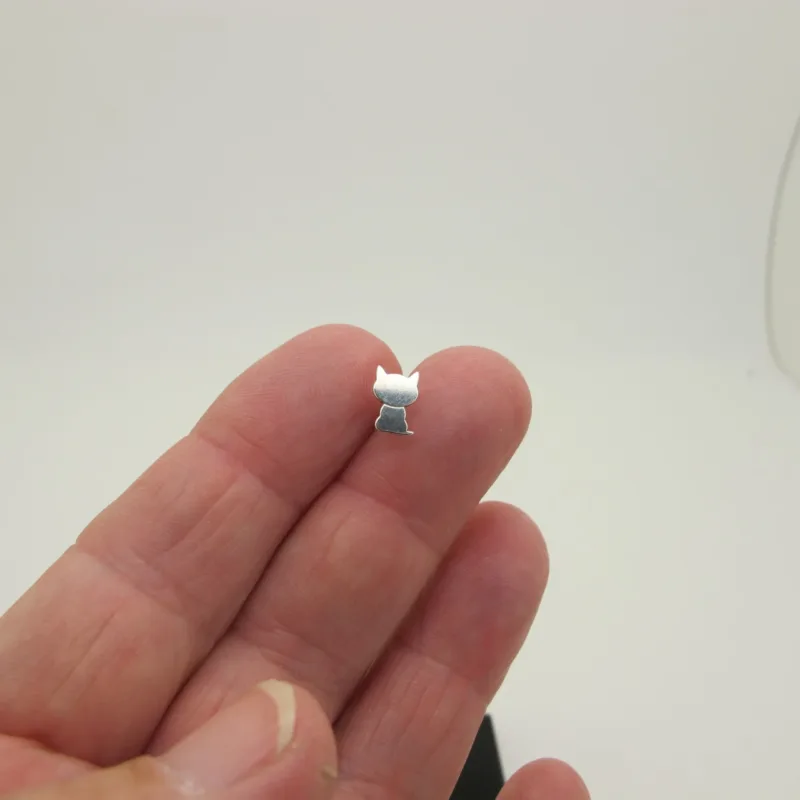En hand håller Örhänge med stift - Katt, ett litet kattformat örhänge i metall med svansen uppåt, mot en vit bakgrund.