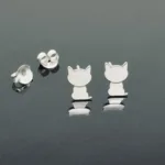 Ett par Örhänge med stift - Katt silverörhängen i form av sittande katter, utställda på en reflekterande mörk yta med två örhängesbrickor bredvid.
