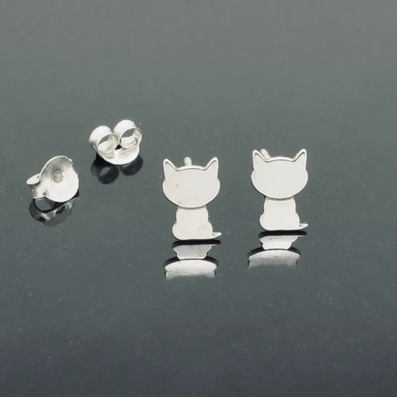 Ett par Örhänge med stift - Katt silverörhängen i form av sittande katter, utställda på en reflekterande mörk yta med två örhängesbrickor bredvid.