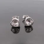 Örhänge med stift Kristallhjärta: Silverfärgade hjärtformade örhängen med klara kristaller, visade på en reflekterande svart yta.