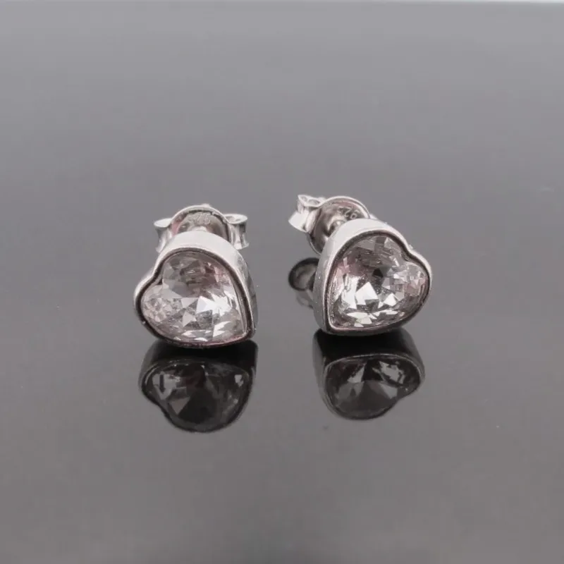 Örhänge med stift Kristallhjärta: Silverfärgade hjärtformade örhängen med klara kristaller, visade på en reflekterande svart yta.