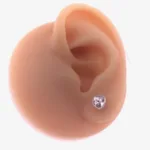 Örhänge med stift Kristallhjärta, ett hjärtformat stiftörhänge i klar kristall, visas på örsnibben på en realistisk köttfärgad öronprotes mot en vit bakgrund.