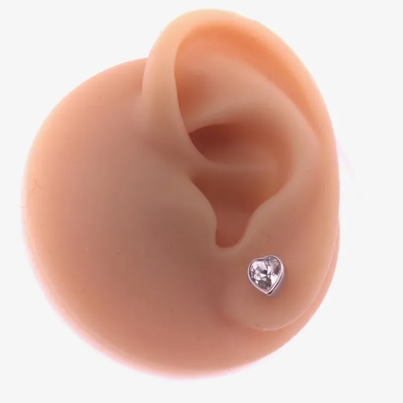 Örhänge med stift Kristallhjärta, ett hjärtformat stiftörhänge i klar kristall, visas på örsnibben på en realistisk köttfärgad öronprotes mot en vit bakgrund.