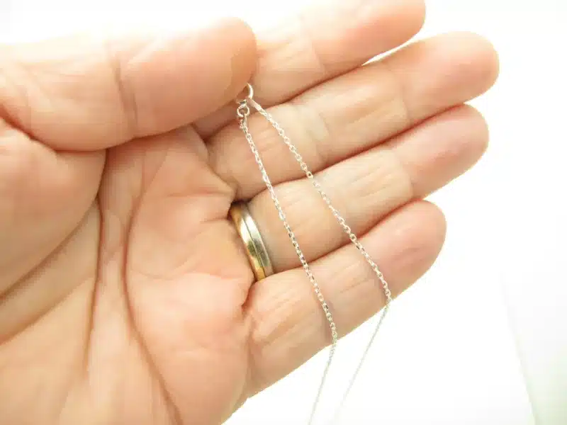 En hand med en guldring på ringfingret håller i Ankarkedja 1,2mm Halskedja 38-50cm silverhalsband mot en vit bakgrund.