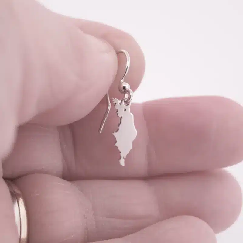 En hand håller i Örhänge Gotland i sterlingsilver, ett litet örhänge format som en ö, med en mjukt suddig bakgrund.