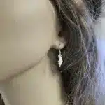 Närbild av en kvinnas öra med Örhänge Gotland i sterlingsilver, ett örhänge format som Gotlands konturer, med löst brunt hår som ramar in hennes ansikte.