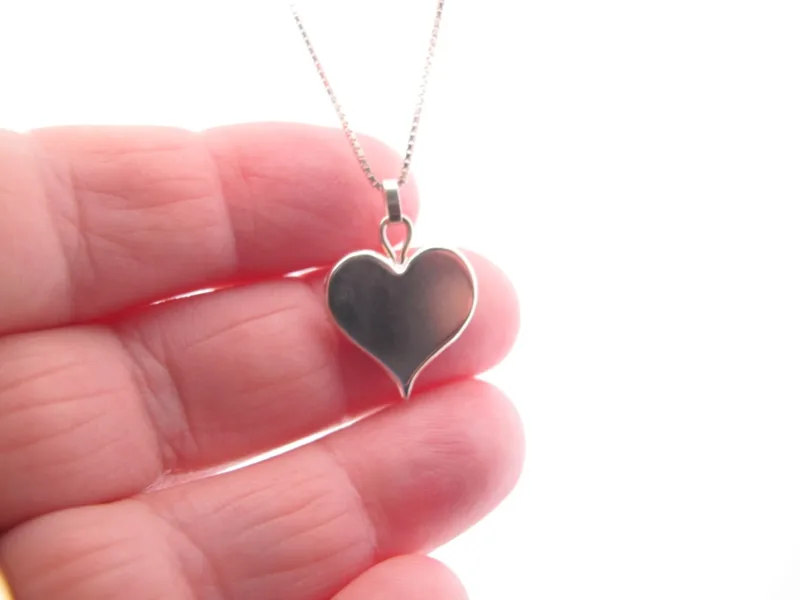 Närbild av en hand som håller i hänget Handgjort Silverhjärta 27x15mm på en fin kedja mot en vit bakgrund.