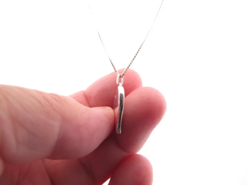 En hand håller i Handgjort Silverhjärta 27x15mm, ett delikat silverhalsband med ett tunt, vertikalt hjärtformat hänge, mot en vit bakgrund.