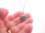 En hand håller Handgjort Silverhjärta 27x15mm hänge på en kedja, som visar dess hjärtform och delikata graverade bokstäver mot en vit bakgrund.