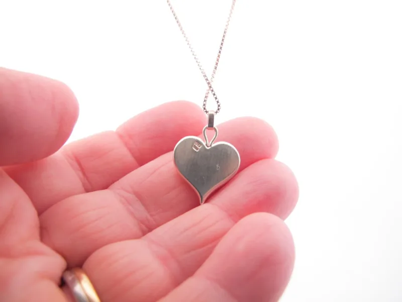 En hand håller Handgjort Silverhjärta 27x15mm hänge på en kedja, som visar dess hjärtform och delikata graverade bokstäver mot en vit bakgrund.