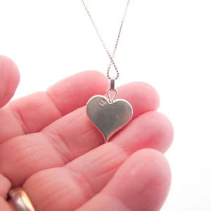 En hand håller Handgjort Silverhjärta 27x15mm hänge på en kedja, som visar dess hjärtform och delikata graverade bokstäver mot en vit bakgrund.