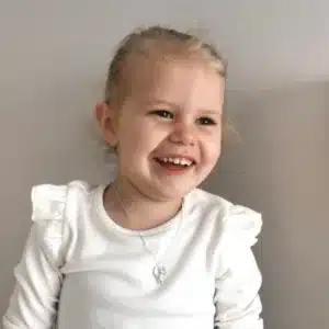 Ett litet barn med ljust hår uppsatt och en vit ruffig långärmad skjorta ler och visar upp Handgjord Silverängel 25mm halsbandet medan hon tittar åt sidan mot en grå bakgrund.
