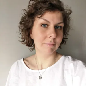 En person med kort, lockigt brunt hår och blå ögon bär en vit topp och visar upp halsbandet Handgjord Silverängel 26mm medan hon poserar mot en grå bakgrund.