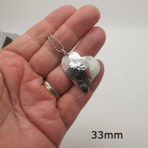 En hand håller i Handgjort Silverhjärta 33x27mm, ett hjärtformat hänge i hamrat silver på en liten kulkedja. Hänget mäter 33mm, vilket framgår av text i nedre högra hörnet.