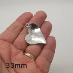 En hand håller i Handgjort Silverhjärta 33x27mm, ett silverglänsande hjärtformat hänge med kedja. "33mm" visas i svart text längst ner till vänster i bilden.