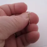 Närbild av en hand som håller i Silverberlock Kvinnosymbol, en tunn metallnål med en liten ögla i ena änden, mot en vit bakgrund.