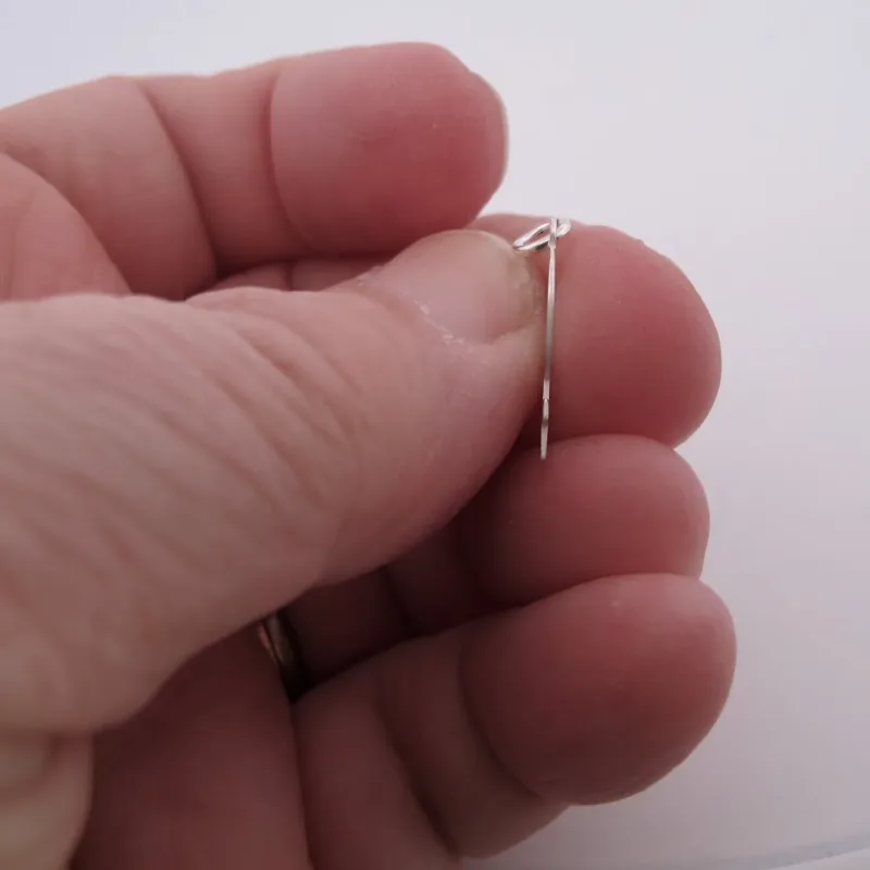 Närbild av en hand som håller i Silverberlock Kvinnosymbol, en tunn metallnål med en liten ögla i ena änden, mot en vit bakgrund.