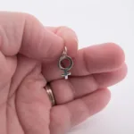 En hand med en vigselring håller Silverberlock Kvinnosymbol, ett litet silverhänge format som den kvinnliga könssymbolen, mot en vit bakgrund.
