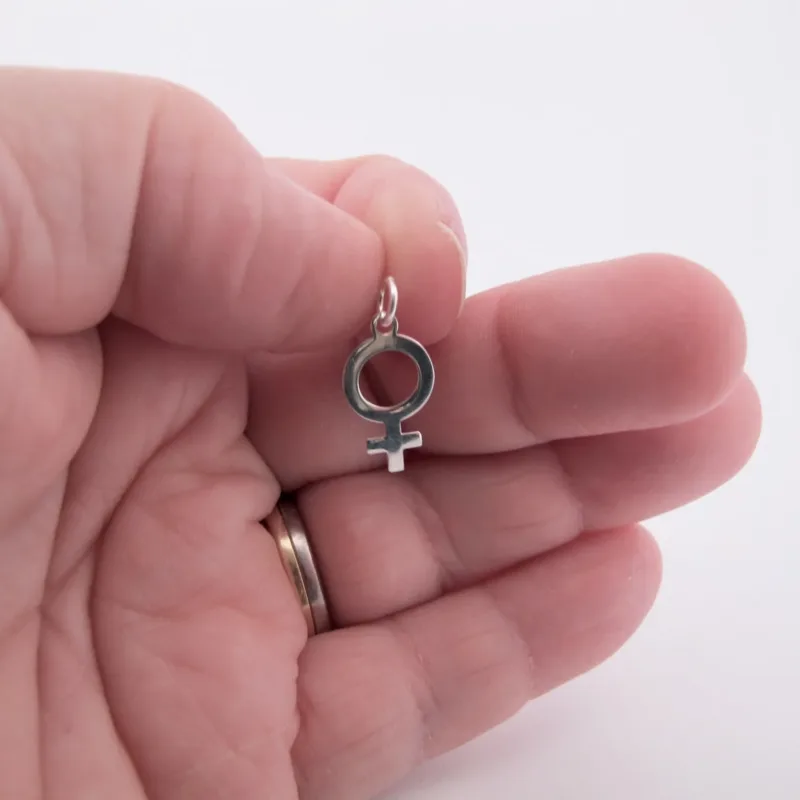 En hand med en vigselring håller Silverberlock Kvinnosymbol, ett litet silverhänge format som den kvinnliga könssymbolen, mot en vit bakgrund.