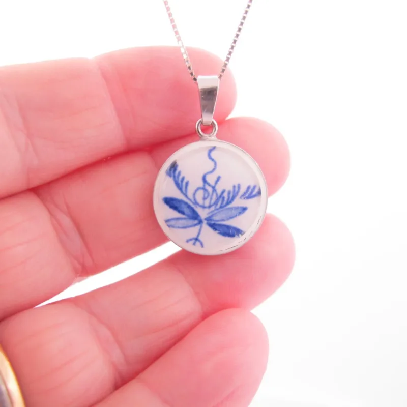 En hand håller i Porslinssmycke - Dresden 25x19mm hängsmycke, med en blå botanisk eller abstrakt design på en vit bakgrund, infattad i en silverram.