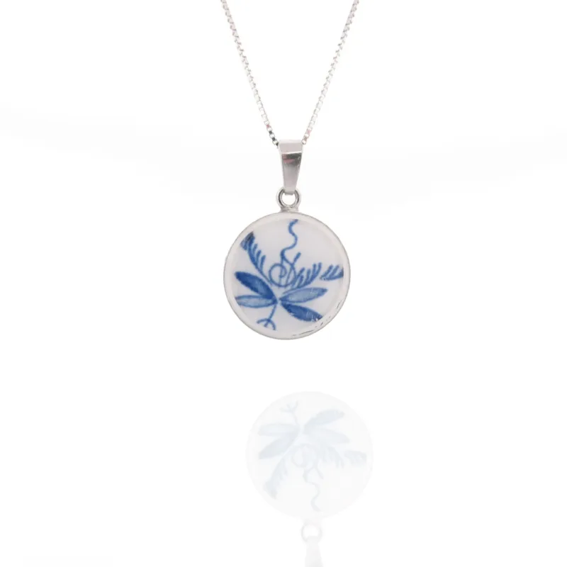 Halsbandet Porslinssmycke - Dresden 25x19mm har ett runt vitt keramiskt hänge med en blå blommig design på en silverkedja, som visas med sin reflektion på en blank yta.