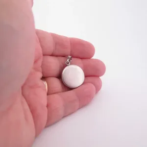 En hand håller i Porslinsmycke - Dresden 20mm, en rund silvermedaljong med slät yta och liten ögla för kedja, mot en vit bakgrund.