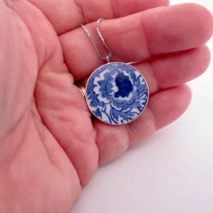 En hand håller i hängsmycket "Porslinssmycke - Gustavsberg 24mm", med en blå och vit blommig porslinsliknande design på en silverkedja.
