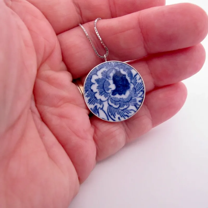 En hand håller i hängsmycket "Porslinssmycke - Gustavsberg 24mm", med en blå och vit blommig porslinsliknande design på en silverkedja.