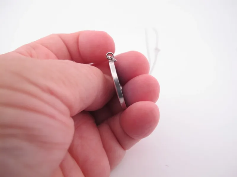 En hand håller i Porslinssmycke - Gustavsberg 24mm, ett tunt silverörhänge, mot en vit bakgrund.