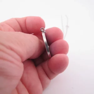 En hand håller i Porslinssmycke - Gustavsberg 24mm, ett tunt silverörhänge, mot en vit bakgrund.