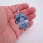 En hand håller i "Porslinssmycke - Gustavsberg"-smycket på en silverkedja, som har en oval design med en blåvit bild av en byggnad med ett torn bland träd.