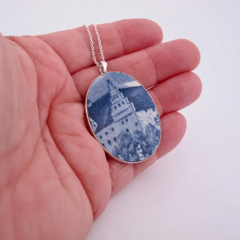 En hand håller i "Porslinssmycke - Gustavsberg"-smycket på en silverkedja, som har en oval design med en blåvit bild av en byggnad med ett torn bland träd.