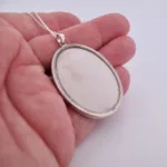 En hand håller i Porslinsmycke - Gustavsberg, ett ovalt silverhänge med slät vit mitt på en tunn silverkedja mot en ljus bakgrund.