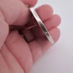 En hand håller Porslinsmycke - Gustavsberg, ett blankt, rektangulärt silverhänge, i änden för att visa dess sidoprofil mot en vit bakgrund.