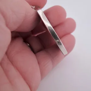 En hand håller Porslinsmycke - Gustavsberg, ett blankt, rektangulärt silverhänge, i änden för att visa dess sidoprofil mot en vit bakgrund.